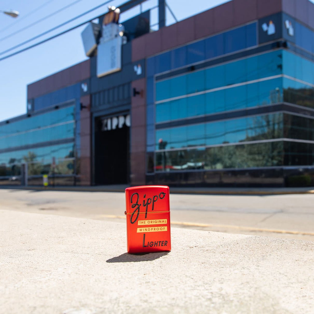 Lifestyle image of Zippo Red Box Top Design Metallic Red Windproof Lighter standing on the sidewalk, with the Zippo headquarters in the background