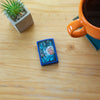 Lifestyle image of Retro Futuristic Royal Blue Matte Windproof Lighter laying on a end table with a plant and a cup of coffee