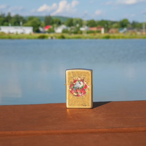 Lifestyle image of Spazuk Bird and Roses Design Brushed Brass Windproof Lighter standing on a railing with a lake behind.