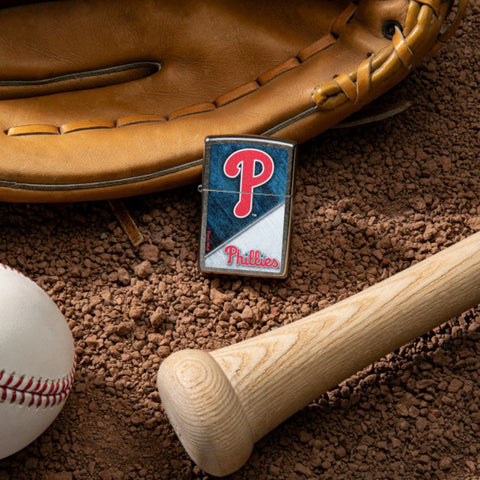 Lifestyle image of MLB® Philadelphia Phillies™ Street Chrome™ Windproof Lighter laying on a baseball field with a glove, ball, and bat.