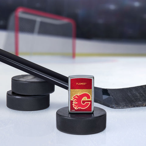 Lifestyle image of the NHL® Calgary Flames Street Chrome™ Windproof Lighter standing with a hockey puck and hockey stick, with a hockey net in the background.