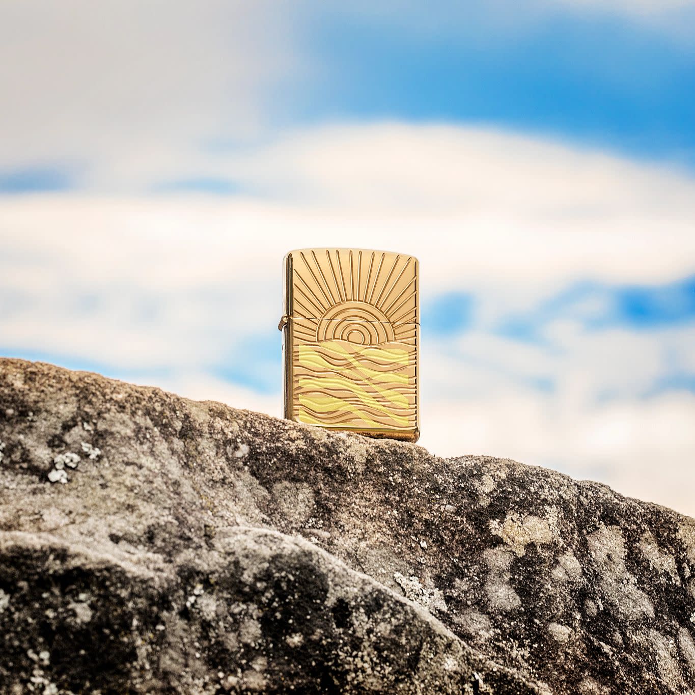Lifestyle image of SunBeam Pendant ArmorÂ® High Polish Brass Windproof Lighter standing on a rock with clouds in the background.