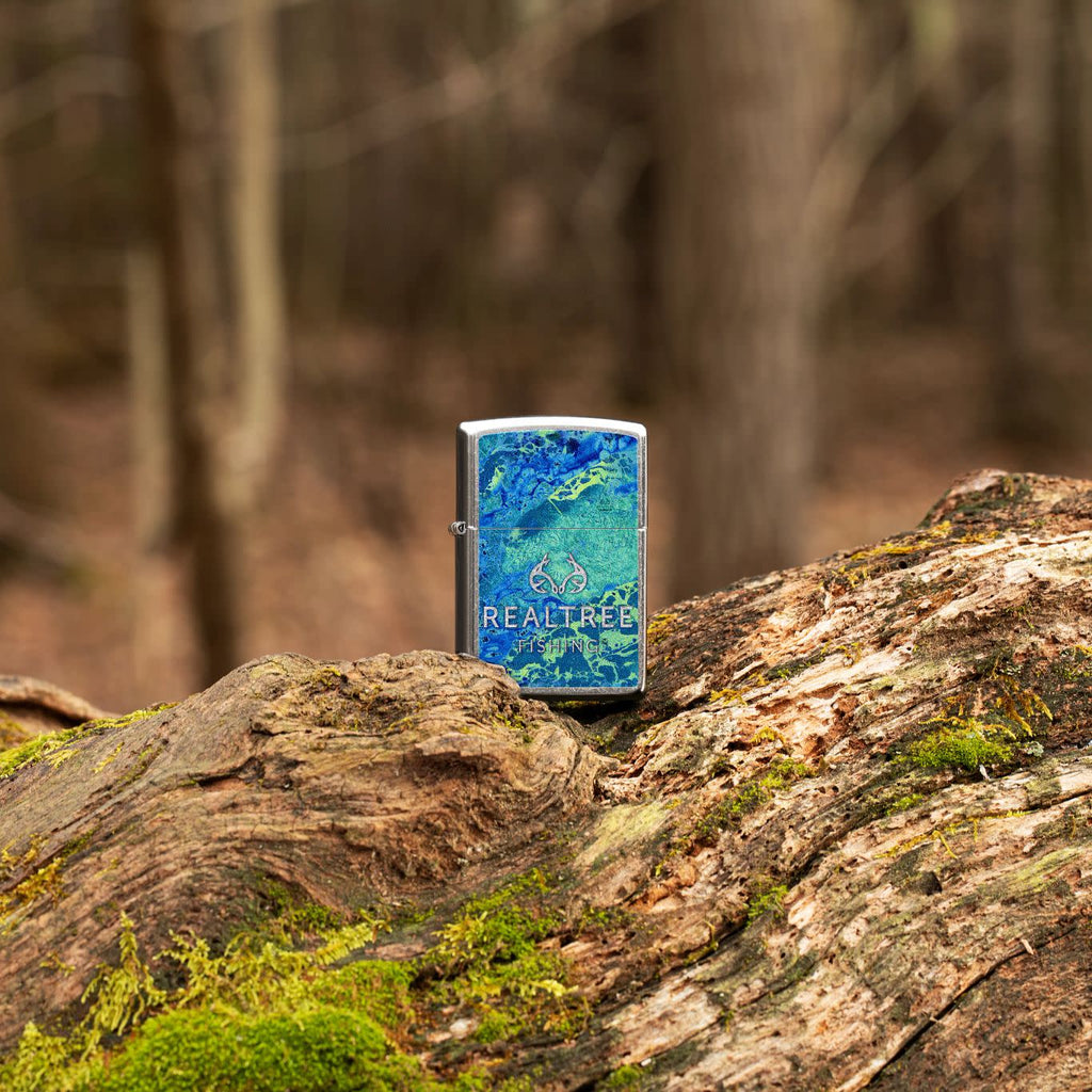 Lifestyle image of Realtree® Blue and Green Camo Street Chrome™ Windproof Lighter standing on a log in the woods.