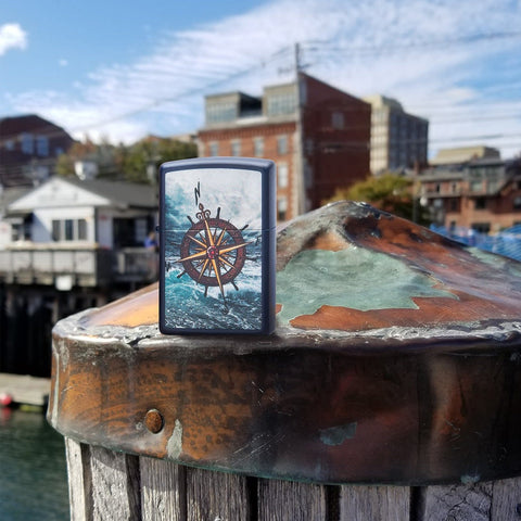 Lifestyle image of Compass Design Navy Matte Windproof Lighter standing on a dock post, with a town in the background.