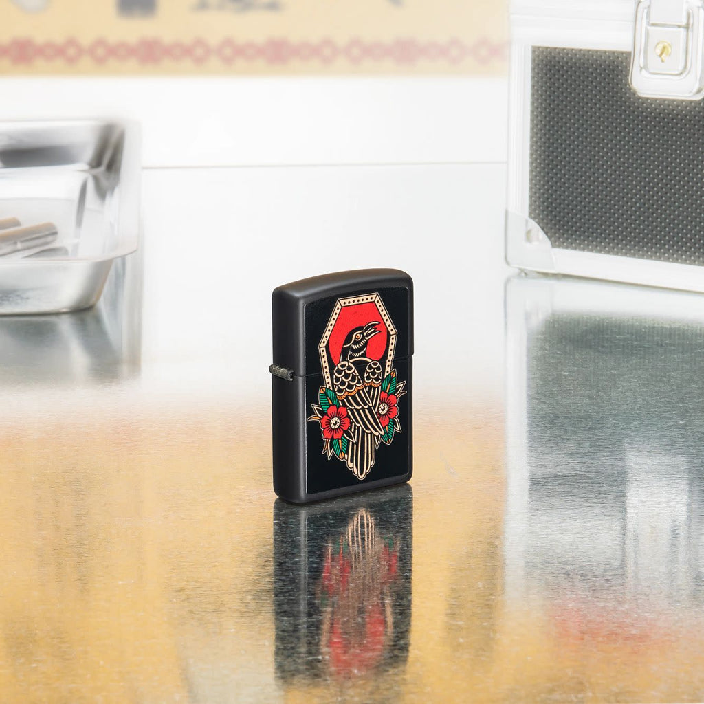 Lifestyle shot of Zippo Crow Tattoo Design Black Matte Windproof Lighter standing in on a reflective table with tattoo equipment.