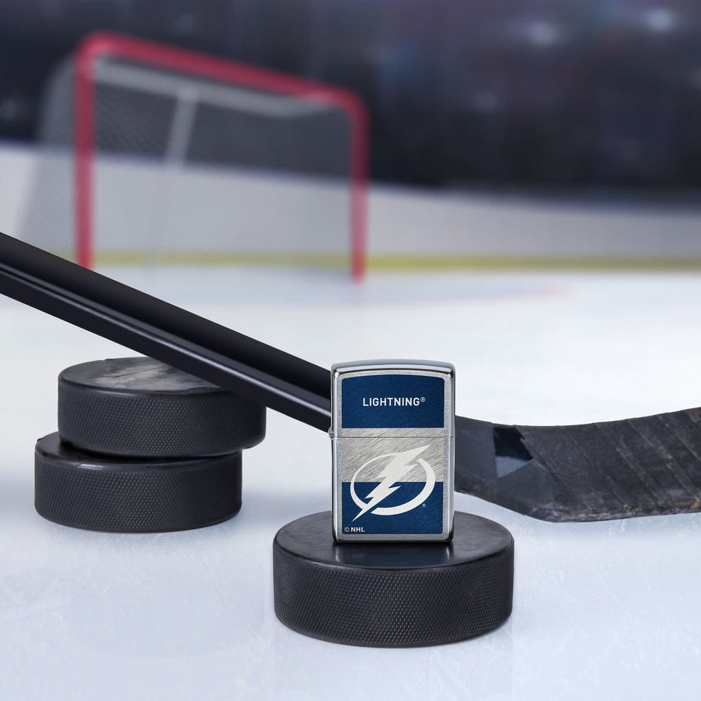 Lifestyle image of the NHL® Tampa Bay Lightning™ Street Chrome™ Windproof Lighter standing with a hockey puck and hockey stick, with a hockey net in the background.