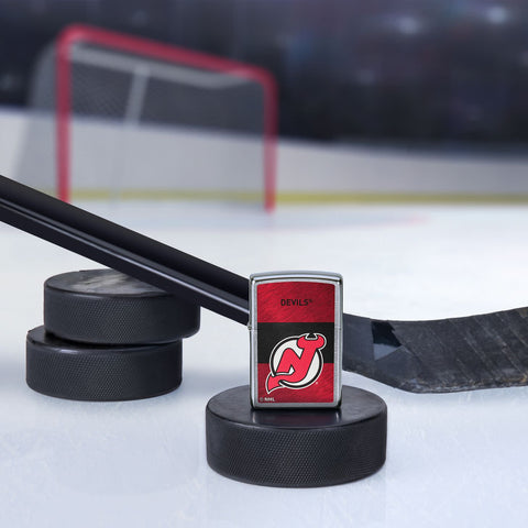 Lifestyle image of the NHL® New Jersey Devils™ Street Chrome™ Windproof Lighter standing with a hockey puck and hockey stick, with a hockey net in the background.