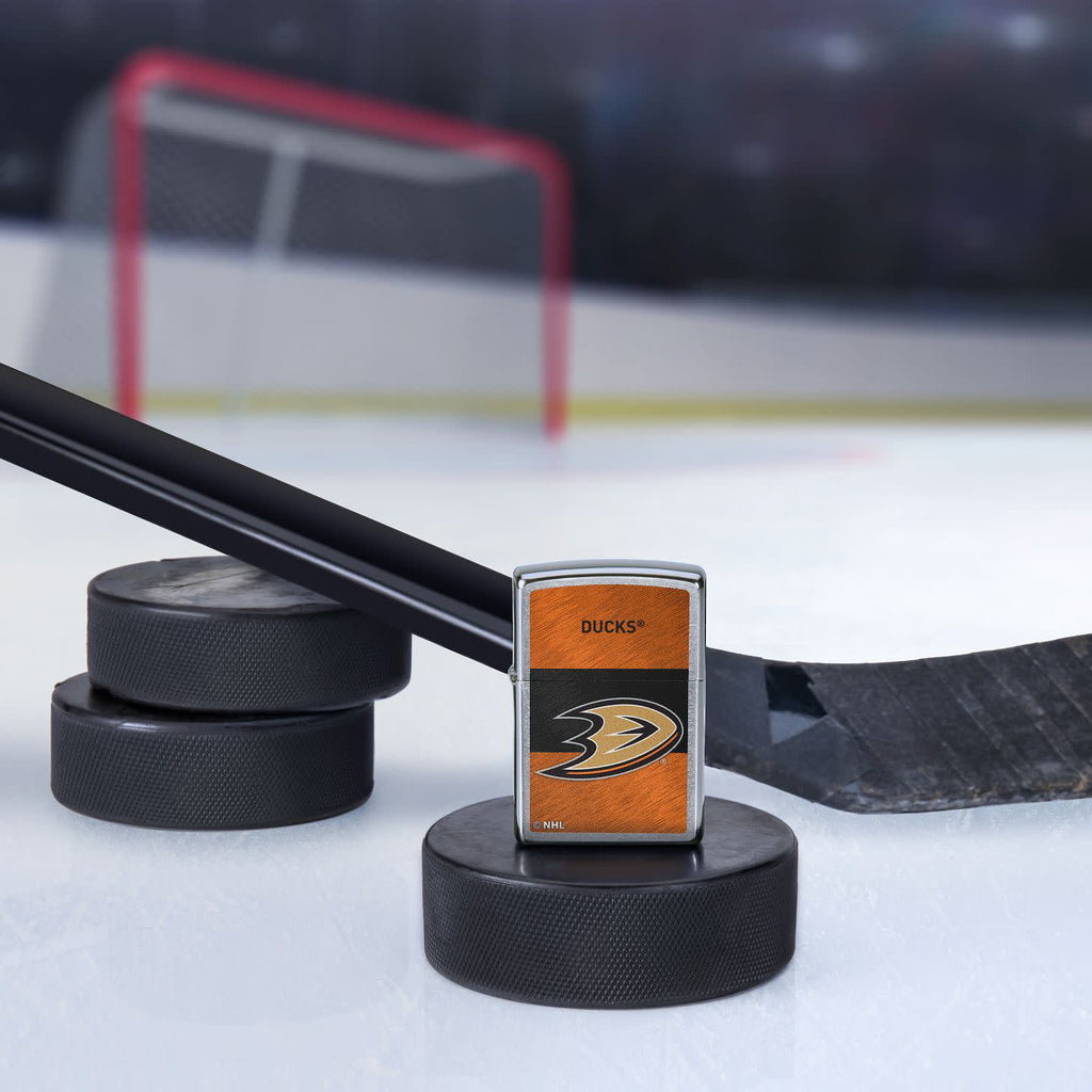 Lifestyle image of the NHL® Anaheim Ducks Street Chrome™ Windproof Lighter standing with a hockey puck and hockey stick, with a hockey net in the background.
