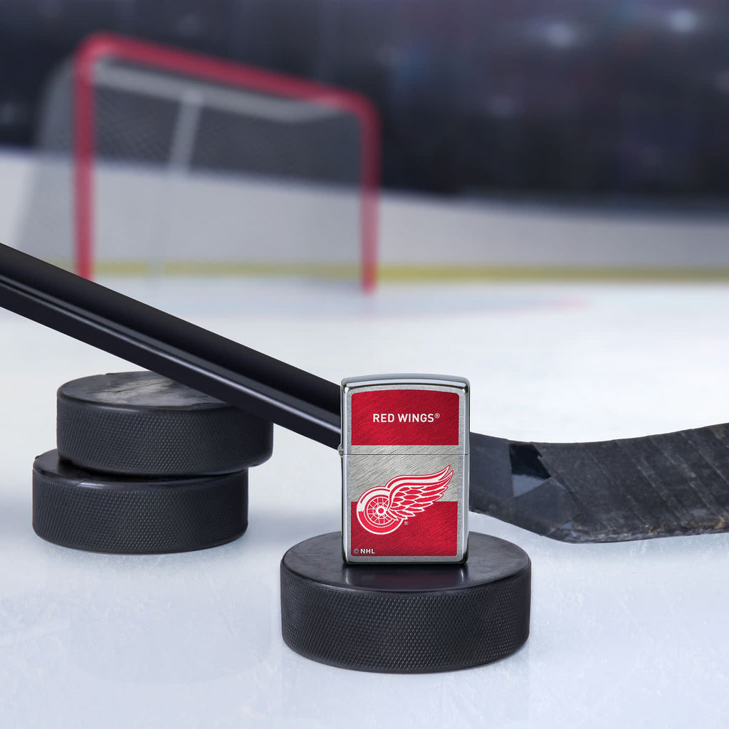 Lifestyle image of the NHL® Detroit Red Wings™ Street Chrome™ Windproof Lighter standing with a hockey puck and hockey stick, with a hockey net in the background.