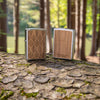 Lifestyle image of two WOODCHUCK USA Walnut Leaves Two-Sided Emblem Windproof Lighters standing on a log. One lighter is showing the front of the design with the other showing the back.