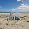 Lifestyle image of two Zippo Guy Harvey 2023 Artist Livestream Windproof Lighters standing on a beach with the ocean in the background.