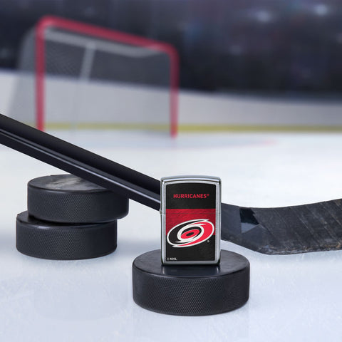 Lifestyle image of the NHL® Carolina Hurricanes™ Street Chrome™ Windproof Lighter standing with a hockey puck and hockey stick, with a hockey net in the background.
