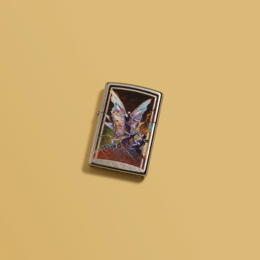 Lifestyle image of Frank Frazetta Mythical Fairy Street Chrome Windproof Lighter laying on a yellow background.