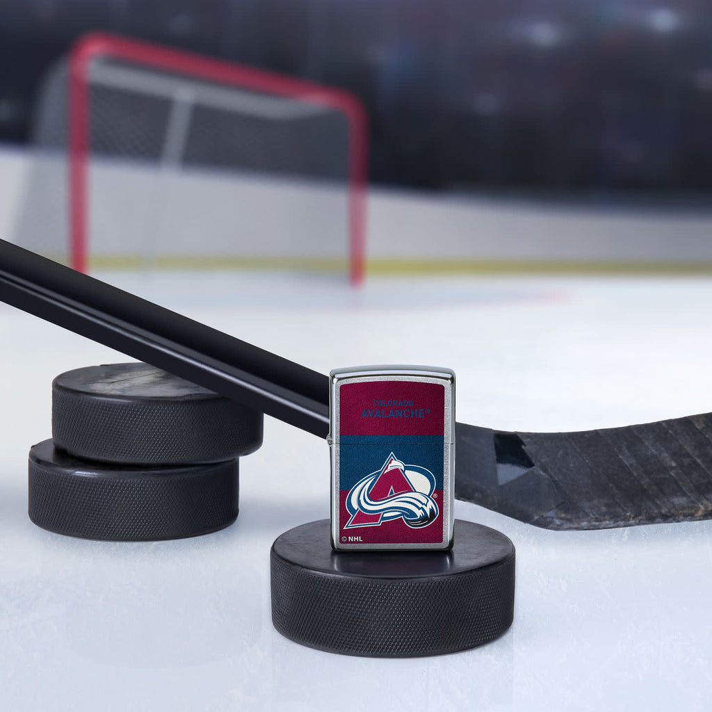 Lifestyle image of the NHL® Colorado Avalanche™ Street Chrome™ Windproof Lighter standing with a hockey puck and hockey stick, with a hockey net in the background.
