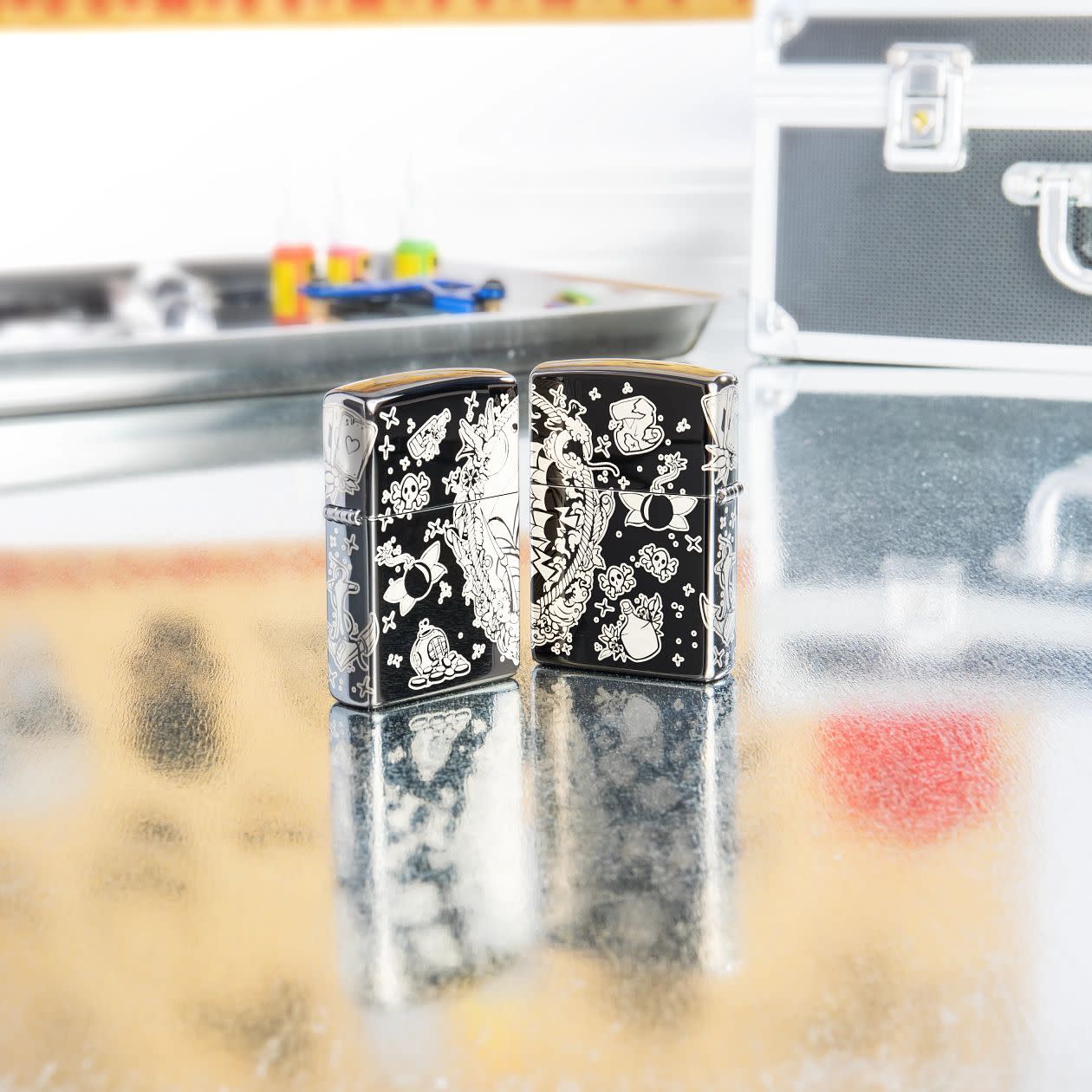 Lifestyle image of two Laser 360Â° Pirates Treasure Design High Polish Black Windproof Lighters standing on a table with tattoo equipment.